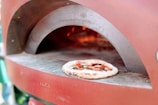 A wood-fired pizza oven with a pizza baking inside. The oven has a rustic appearance with its arched opening, and the flames from the fire are visible in the background. The pizza is topped with tomatoes, basil, and other ingredients, and is placed on a stone surface inside the oven.