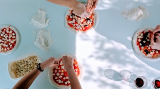 Several hands are seen preparing pizzas on a light blue surface. The pizzas have a base of tomato sauce and are being topped with cubes of cheese and olives. Flour is scattered on the table, and a container filled with cheese cubes is visible. Two glasses of red wine are also present, casting shadows on the surface.