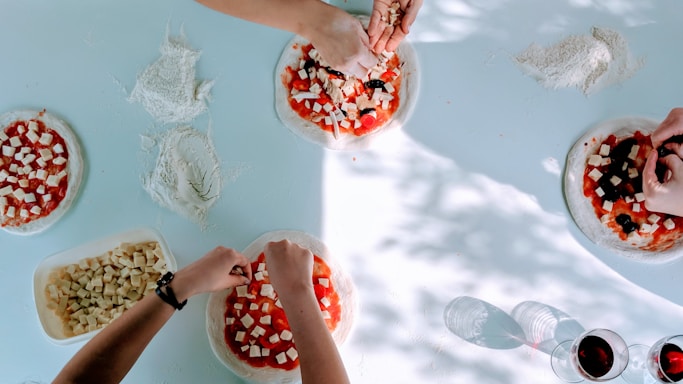 Several hands are seen preparing pizzas on a light blue surface. The pizzas have a base of tomato sauce and are being topped with cubes of cheese and olives. Flour is scattered on the table, and a container filled with cheese cubes is visible. Two glasses of red wine are also present, casting shadows on the surface.