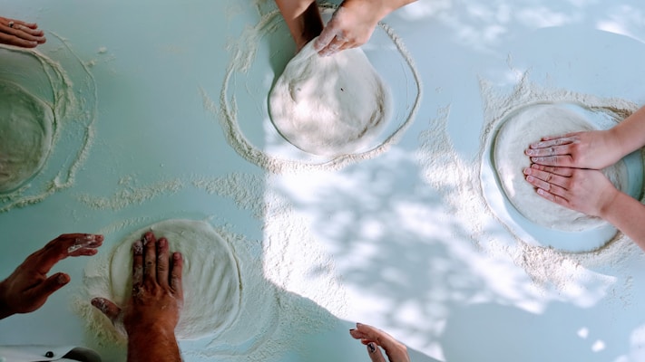 Several pairs of hands are shaping rounds of dough on a floured surface, with sunlight casting dappled shadows over the scene. The hands appear to be engaged in a communal baking activity, suggesting teamwork and cooperation.