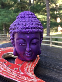 A small purple Buddha head sculpture rests on a decorative red and gold object set on a wooden surface. The background features a blurred view of green trees and a wooden fence, suggesting an outdoor setting.