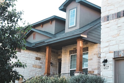 A modern house exterior featuring stone and wood materials. The house has a gabled roof and a prominent window on the upper level. There is greenery in the form of bushes and trees around the house.