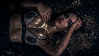 A person lies on a dark surface with golden glitter scattered across their skin and hair. The lighting creates a dramatic contrast, highlighting the glitter and the person's features. They wear a decorated garment, and their expression is serene yet powerful.