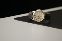 A wristwatch with a brown leather strap is placed on a two-toned surface, partially black and partially white. The watch features a white dial with Roman numeral hour markers and multiple sub-dials, giving it a classic and elegant appearance.