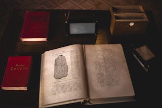 An arrangement of vintage and antique books on a dark wooden table, with an open book displaying illustrations and text. Nearby are other closed books, a wooden box with cards, and a small black box.