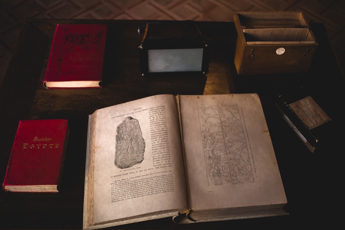 An arrangement of vintage and antique books on a dark wooden table, with an open book displaying illustrations and text. Nearby are other closed books, a wooden box with cards, and a small black box.