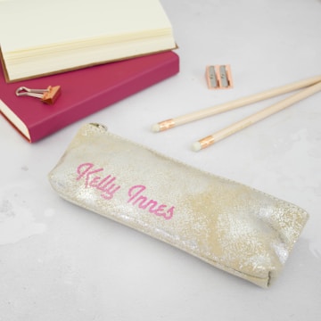 A glittery gold pencil case with the name 'Kelly Innes' written in pink sits on a light gray surface. Nearby, there are pencils with erasers, a double pencil sharpener, and an open notebook with a pink cover. A rose gold binder clip lies next to the notebook.