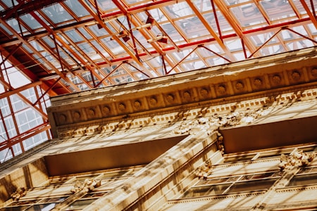 A detailed perspective of an architectural structure with intricate detailing on the facade. Above it, a modern glass and metal ceiling structure creates a stark contrast, casting shadows on the older building. The interplay of light and shadow enhances the texture and depth of the architecture.