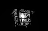 A black and white image of a lantern-style light fixture. The light bulb is visible, emitting a bright glow that highlights the metal bars of the fixture. The background is dark, emphasizing the contrast between the light and the surrounding darkness.