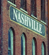 A brick building facade featuring a large sign painted with the word 'Nashville' in bold white letters. The structure has a vintage architectural style, characterized by arched windows and weathered brickwork.