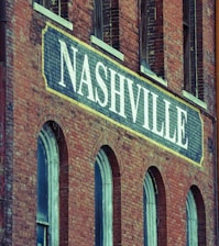A brick building facade featuring a large sign painted with the word 'Nashville' in bold white letters. The structure has a vintage architectural style, characterized by arched windows and weathered brickwork.