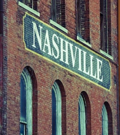 A brick building facade featuring a large sign painted with the word 'Nashville' in bold white letters. The structure has a vintage architectural style, characterized by arched windows and weathered brickwork.