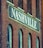 A brick building facade featuring a large sign painted with the word 'Nashville' in bold white letters. The structure has a vintage architectural style, characterized by arched windows and weathered brickwork.