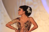 A woman with a side profile is wearing an ornate dress adorned with beads and sequins. Her hair is styled elegantly with an embellished hair accessory shaped like a butterfly. She stands confidently with her hands on her hips against a blurred background.