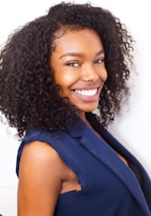 A person with curly hair and a nose piercing is smiling, wearing a dark blue sleeveless top.