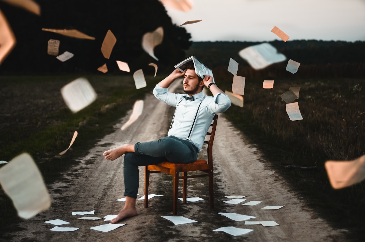 Photograph of a man sitting on a char, in the middle of  a dirt road with a book over his head.