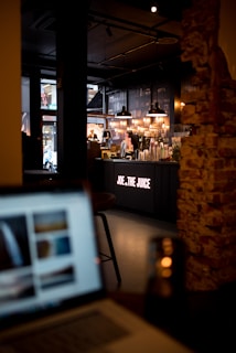 A dimly lit cafe interior with modern industrial decor. The centerpiece is the counter of a juice bar named 'Joe & The Juice' where several drink mixers and equipment are visible. Warm lighting is provided by hanging pendant lights above the counter. In the foreground, there is a blurred image of a laptop, out of focus. Exposed brick walls add to the cozy ambiance.