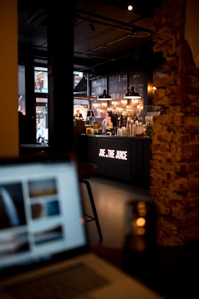 A dimly lit cafe interior with modern industrial decor. The centerpiece is the counter of a juice bar named 'Joe & The Juice' where several drink mixers and equipment are visible. Warm lighting is provided by hanging pendant lights above the counter. In the foreground, there is a blurred image of a laptop, out of focus. Exposed brick walls add to the cozy ambiance.