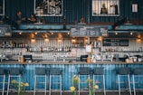 A stylish bar setup within a shipping container features a well-stocked bar with a variety of bottles and glasses on display. The interior has an industrial aesthetic with metal and wood elements. Several bar stools are neatly positioned along the counter. Overhead, there are warm lights creating a cozy atmosphere, and above the bar, there are blackboards displaying the menu. In the upper level, two separate people are engaging in conversations in small seating areas.