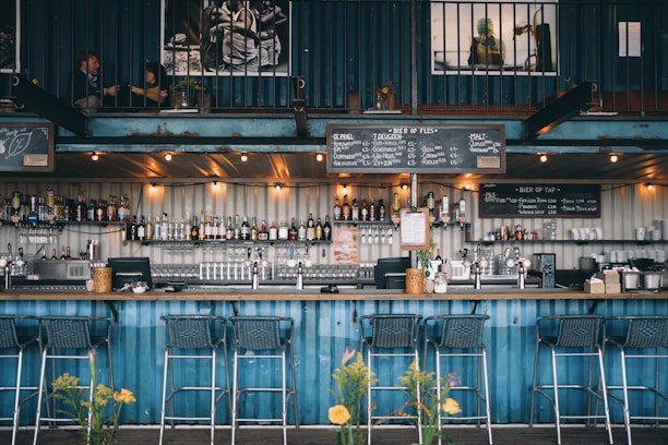 A stylish bar setup within a shipping container features a well-stocked bar with a variety of bottles and glasses on display. The interior has an industrial aesthetic with metal and wood elements. Several bar stools are neatly positioned along the counter. Overhead, there are warm lights creating a cozy atmosphere, and above the bar, there are blackboards displaying the menu. In the upper level, two separate people are engaging in conversations in small seating areas.