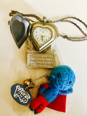 A heart-shaped pocket watch with a chain is lying open, revealing a classic clock face. Next to it, a small blue and red voodoo doll keychain is visible, with a tag that reads 'Whatever Vodka'. Below them is a wooden plaque with the phrase 'Live every moment, laugh every day and love beyond words'.