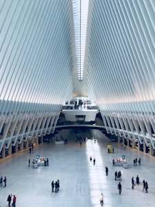 A futuristic and spacious architectural interior characterized by high ceilings and a symmetrical design. The space is lined with white rib-like beams and features a large central atrium with people walking and interacting. The floor is polished and reflective, and the light from the ceiling windows creates patterns of shadows on the floor.