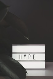 A lightbox with the word 'HYPE' illuminated in bold letters, set against a dark background with large green leaves partially in view.