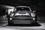 A sleek, modern vehicle parked in a dimly lit, industrial-looking garage. The car has a distinctive design with large, round headlights and a bold front grille. The lighting creates dramatic shadows and highlights on the car's surface, emphasizing its contours.