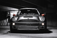 A sleek, modern vehicle parked in a dimly lit, industrial-looking garage. The car has a distinctive design with large, round headlights and a bold front grille. The lighting creates dramatic shadows and highlights on the car's surface, emphasizing its contours.