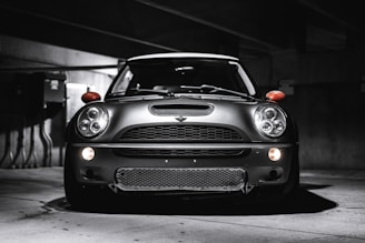 A sleek, modern vehicle parked in a dimly lit, industrial-looking garage. The car has a distinctive design with large, round headlights and a bold front grille. The lighting creates dramatic shadows and highlights on the car's surface, emphasizing its contours.