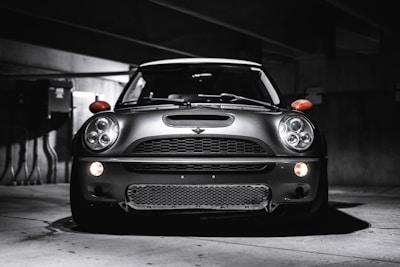 A sleek, modern vehicle parked in a dimly lit, industrial-looking garage. The car has a distinctive design with large, round headlights and a bold front grille. The lighting creates dramatic shadows and highlights on the car's surface, emphasizing its contours.
