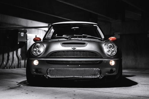 A sleek, modern vehicle parked in a dimly lit, industrial-looking garage. The car has a distinctive design with large, round headlights and a bold front grille. The lighting creates dramatic shadows and highlights on the car's surface, emphasizing its contours.