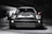 A sleek, modern vehicle parked in a dimly lit, industrial-looking garage. The car has a distinctive design with large, round headlights and a bold front grille. The lighting creates dramatic shadows and highlights on the car's surface, emphasizing its contours.