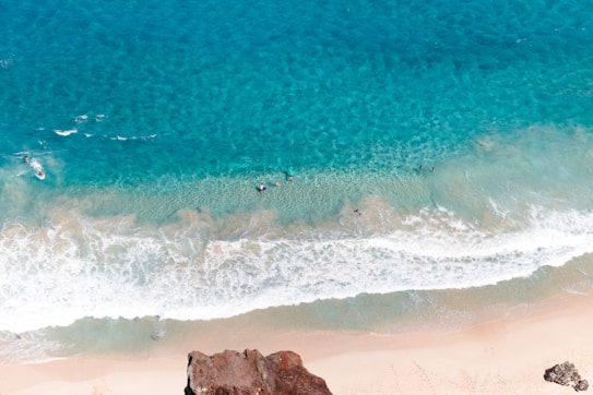 Crystal clear turquoise ocean waves gently lap against a sandy beach, while several people are seen enjoying the water. The perspective appears to be aerial, showing a wide expanse of sea and shoreline. Rocky outcrops frame the scene at the bottom of the image, adding contrast to the bright blue and white hues.