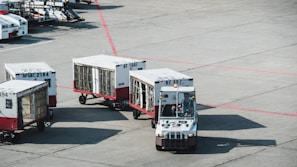 A small airport vehicle is pulling several baggage carts on the tarmac. The number BT 897 is visible on the vehicle, and each cart is labeled with different numbers. The setting appears to be an airport runway area with red lines marking the concrete ground.