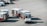 A small airport vehicle is pulling several baggage carts on the tarmac. The number BT 897 is visible on the vehicle, and each cart is labeled with different numbers. The setting appears to be an airport runway area with red lines marking the concrete ground.