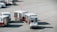 A small airport vehicle is pulling several baggage carts on the tarmac. The number BT 897 is visible on the vehicle, and each cart is labeled with different numbers. The setting appears to be an airport runway area with red lines marking the concrete ground.