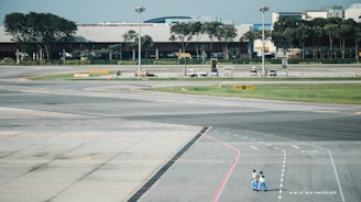 A large airport tarmac with marked lanes is visible in the foreground. There is an open space with concrete pavement. Two people wearing blue uniforms walk across the tarmac. In the background, there are airport structures, vehicles, and equipment, along with a row of trees lining the side. The scene is well-lit, suggesting daylight conditions.