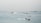 Multiple boats and ships are scattered across a large body of water, with several ferries visible in the foreground creating trails in the calm sea. The background shows a horizon line filled with additional vessels, indicating busy maritime activity under a hazy sky.