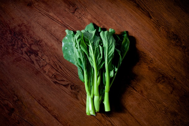 A bunch of fresh green leafy vegetables is placed on a rustic wooden surface. The leaves are vibrant and healthy, with dark green hues and crisp texture.