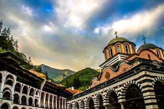 A picturesque view of a colorful, ornate building with curved domes and intricate patterns, set against a backdrop of lush green mountains and a dramatic sky filled with clouds. The architecture combines elements typical of Eastern Orthodox Christian monasteries, featuring arches and striped decorations.