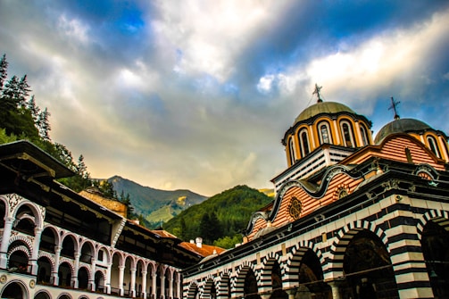 A picturesque view of a colorful, ornate building with curved domes and intricate patterns, set against a backdrop of lush green mountains and a dramatic sky filled with clouds. The architecture combines elements typical of Eastern Orthodox Christian monasteries, featuring arches and striped decorations.