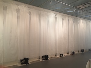 A row of vintage sewing machines is lined up against a backdrop of sheer white curtains, hanging from the ceiling, creating an atmosphere reminiscent of an art installation. The machines are placed on the floor at regular intervals, and the entire scene is set in a minimalist, modern space with a reflective ceiling.