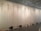 A row of vintage sewing machines is lined up against a backdrop of sheer white curtains, hanging from the ceiling, creating an atmosphere reminiscent of an art installation. The machines are placed on the floor at regular intervals, and the entire scene is set in a minimalist, modern space with a reflective ceiling.