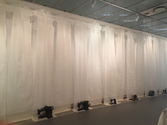 A row of vintage sewing machines is lined up against a backdrop of sheer white curtains, hanging from the ceiling, creating an atmosphere reminiscent of an art installation. The machines are placed on the floor at regular intervals, and the entire scene is set in a minimalist, modern space with a reflective ceiling.