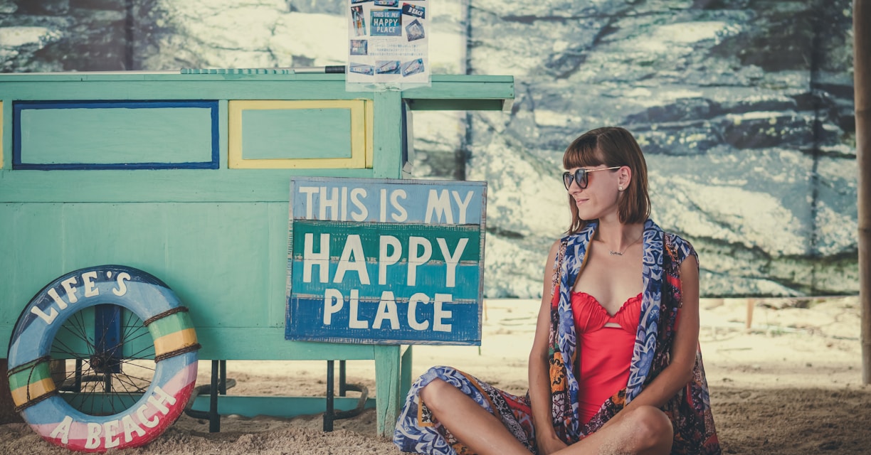 woman sitting on sand beside of This is My Happy Place signage