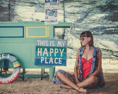 woman sitting on sand beside of This is My Happy Place signage