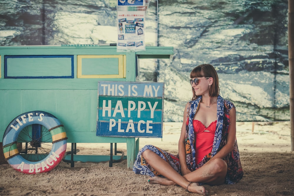 mujer sentada en la arena al lado de la señalización de This is My Happy Place
