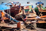 Several wooden boats with painted hulls are docked at a harbor. The boats have blue canvas canopies and are tied with thick ropes to the pier. Some boats display registration numbers on their sides, and various items are visible on board, including packages and colorful fabrics.