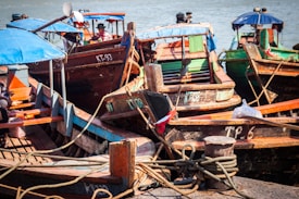 Several wooden boats with painted hulls are docked at a harbor. The boats have blue canvas canopies and are tied with thick ropes to the pier. Some boats display registration numbers on their sides, and various items are visible on board, including packages and colorful fabrics.